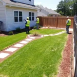 A residential house with a newly landscaped yard: a tidy lawn, a small flower bed with colorful plants, a stone pathway leading to the front door, a wooden fence, and a few construction workers in safety vests using tools to repair the surrounding area. The scene shows a clean, organized environment with subtle landscaping details and a bright, sunny sky.