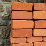 A close-up of a stack of handmade clay bricks, each with a slightly uneven, earthy texture, arranged in a way that suggests they are being laid for a traditional stove. The bricks should be warm-toned, with subtle variations in color from light brown to deep reddish-brown, and a faint glaze sheen. The background should be a muted, rustic stone wall, with soft natural lighting highlighting the bricks' surface details.