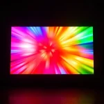 A vibrant, high-definition TV screen displaying a dynamic burst of rich, saturated colors — deep crimson, electric blue, luminous gold, and emerald green — radiating outward in smooth gradients and soft glows, as if the screen is glowing from within. The surrounding environment is dark and minimalistic, emphasizing the screen’s luminosity. No text, logos, or UI elements visible on the screen. The image conveys energy, clarity, and visual brilliance, capturing the essence of a TV screen 'shining