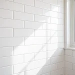 A close-up view of a modern bathroom wall covered in elegant white ceramic tiles with subtle gray grout, arranged in a uniform grid pattern. Soft natural light from a nearby window highlights the clean, minimalist aesthetic, creating a serene and inviting atmosphere.